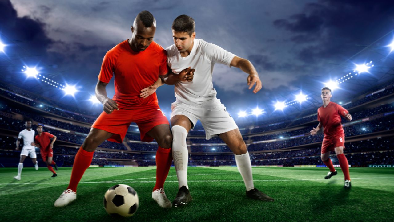 Ein Fußballspieler dribbelt den Ball auf einem Fußballplatz in einem flutlichtbeleuchteten Freiluftstadion voller Zuschauer*innen unter einem stürmischen Abendhimmel.