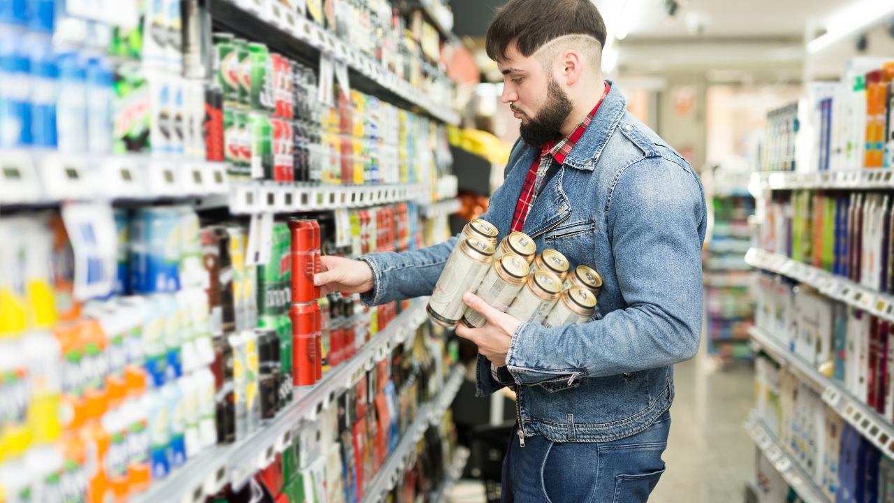Ein junger bärtiger Mann kauft im Supermarkt Getränke ein und nimmt Dosenbier aus den Regalen.