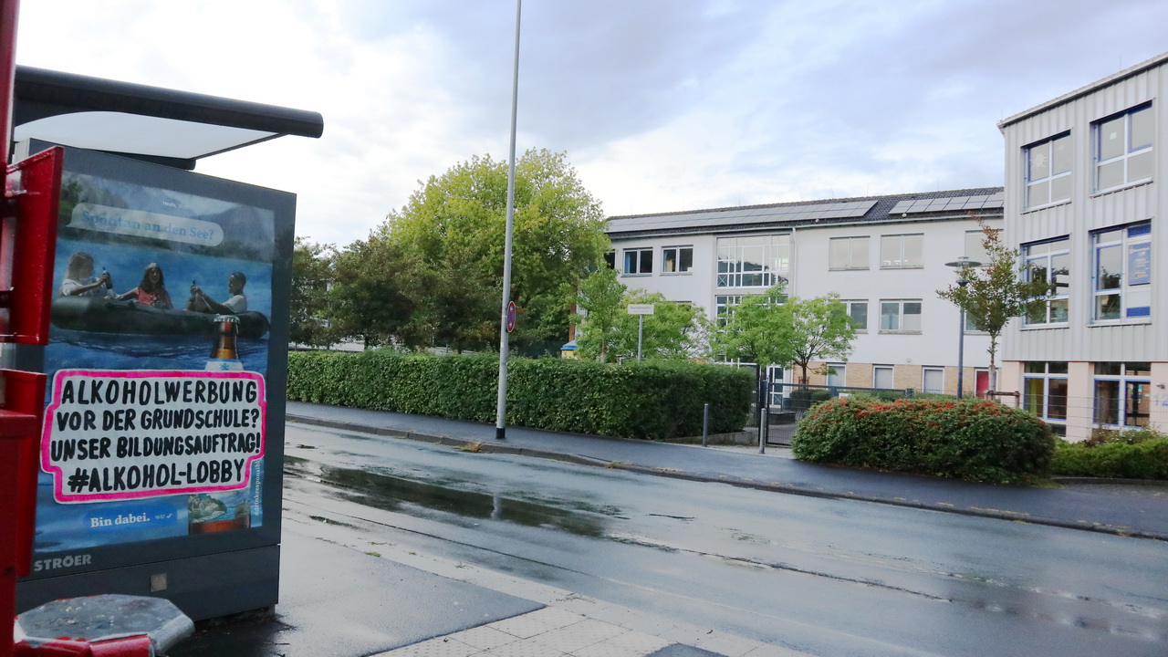 Bushaltestelle mit Bierwerbung vor einem Schulgebäude an einer regennassen Straße. Auf dem Werbeplakat, das drei Personen mit Bierflaschen in einem Schlauchboot zeigt, klebt ein Protestaufkleber mit der Aufschrift: 'Alkoholwerbung vor der Grundschule? Unser Bildungsauftrag! #Alkohol-Lobby'. Im Hintergrund sind ein modernes Schulgebäude, eine Hecke und Bäume unter bewölktem Himmel zu sehen.
