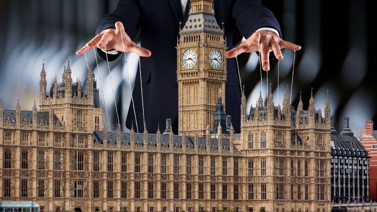 Symbolische Darstellung: Riesige Hände eines Mannes in einem Anzug steuern das britische Parlament (Palace of Westminster) wie eine Marionette an Fäden. Im Hintergrund eine unscharf aufgenommen Reihe von Weinflaschen.