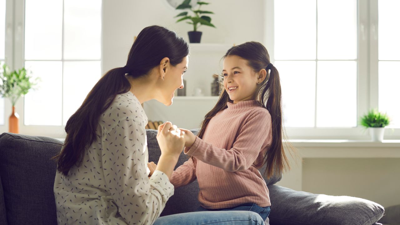 Mutter und Tochter sitzen sich auf einem grauen Sofa gegenüber, halten sich an den Händen und lächeln sich in einem hellen Wohnzimmer an.