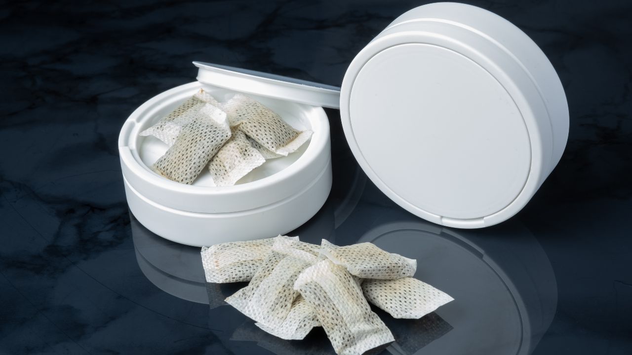 Geöffnete weiße Runddose mit Nicotinbeuteln (Snus-Pouches) darin und einige Beutel davor auf dunkler Marmoroberfläche. Der Deckel steht angelehnt dahinter.