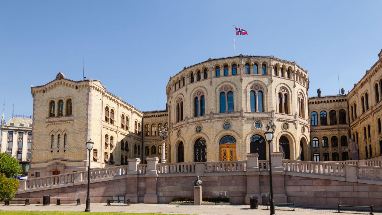 Frontalansicht des Storting, dem Parlamentsgebäude Norwegens in Oslo. Das historische Gebäude aus hellgelbem Backstein zeigt einen charakteristischen halbrunden Mittelbau mit Rundbogenfenstern, flankiert von zwei Seitenflügeln. Auf dem Dach weht die norwegische Flagge vor strahlend blauem Himmel. Vor dem Gebäude befinden sich eine steinerne Balustrade, Laternen und eine Bronzebüste.