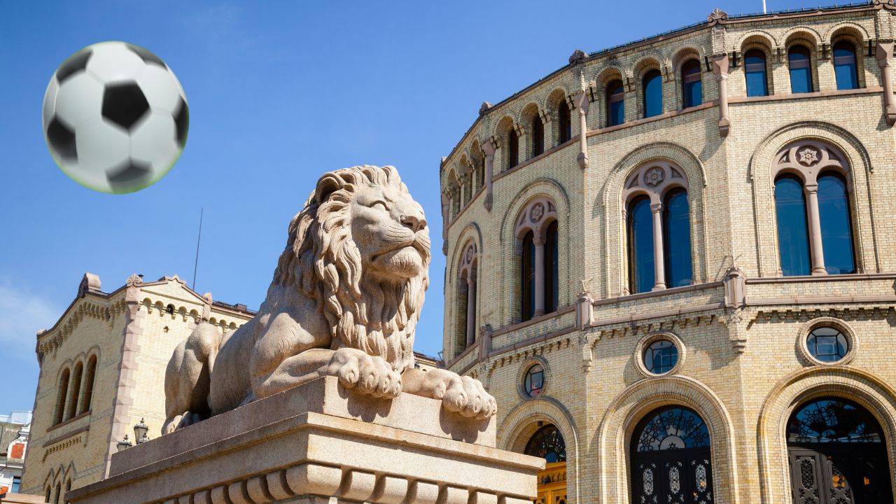Liegende detailreiche Steinlöwen-Statue auf einem Sockel vor dem Storting, dem norwegischen Parlament in Oslo. Aufnahme aus Froschperspektive bei strahlend blauem Himmel, die gelbe Backstein-Fassade des Gebäudes mit neoromanischen Bögen im Hintergrund. Über dem Löwen schwebt ein digital eingefügter schwarz-weißer Fußball in der Luft.