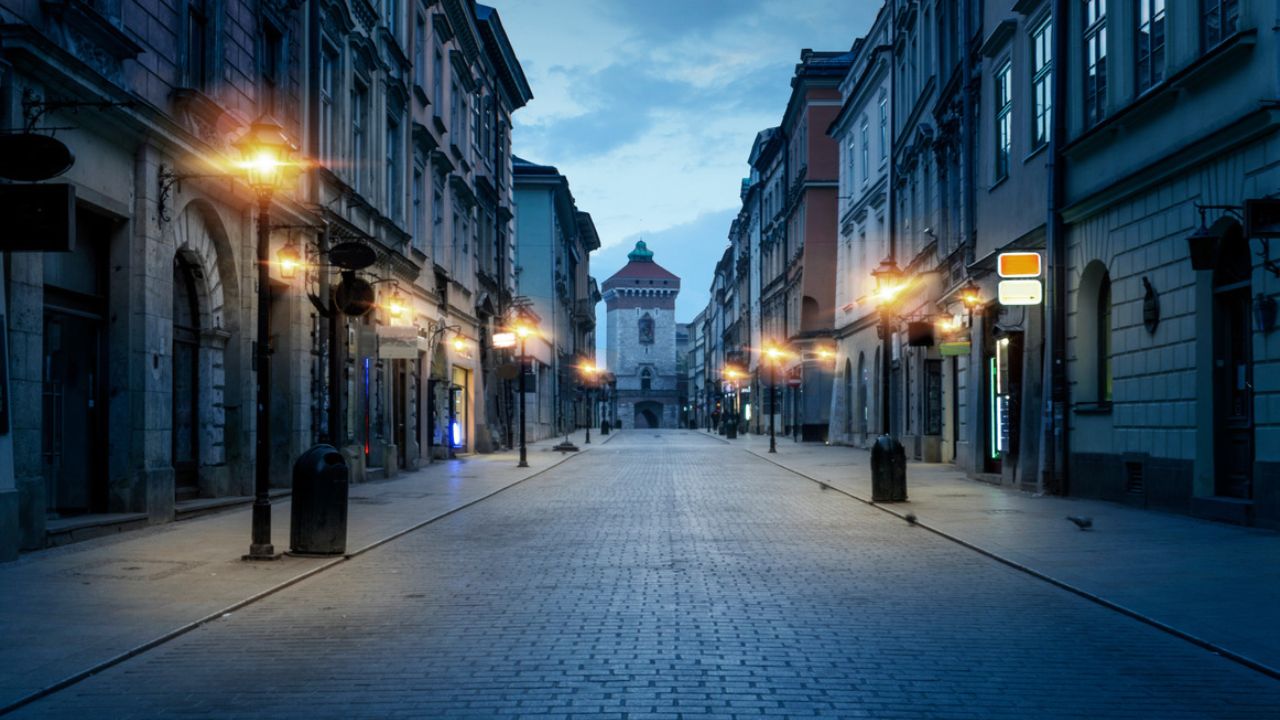 Altstadtstraße bei Dämmerung mit Kopfsteinpflaster, beleuchteten Straßenlaternen und historischen Häusern, Blick auf einen Turm mit grünem Kuppeldach am Straßenende.