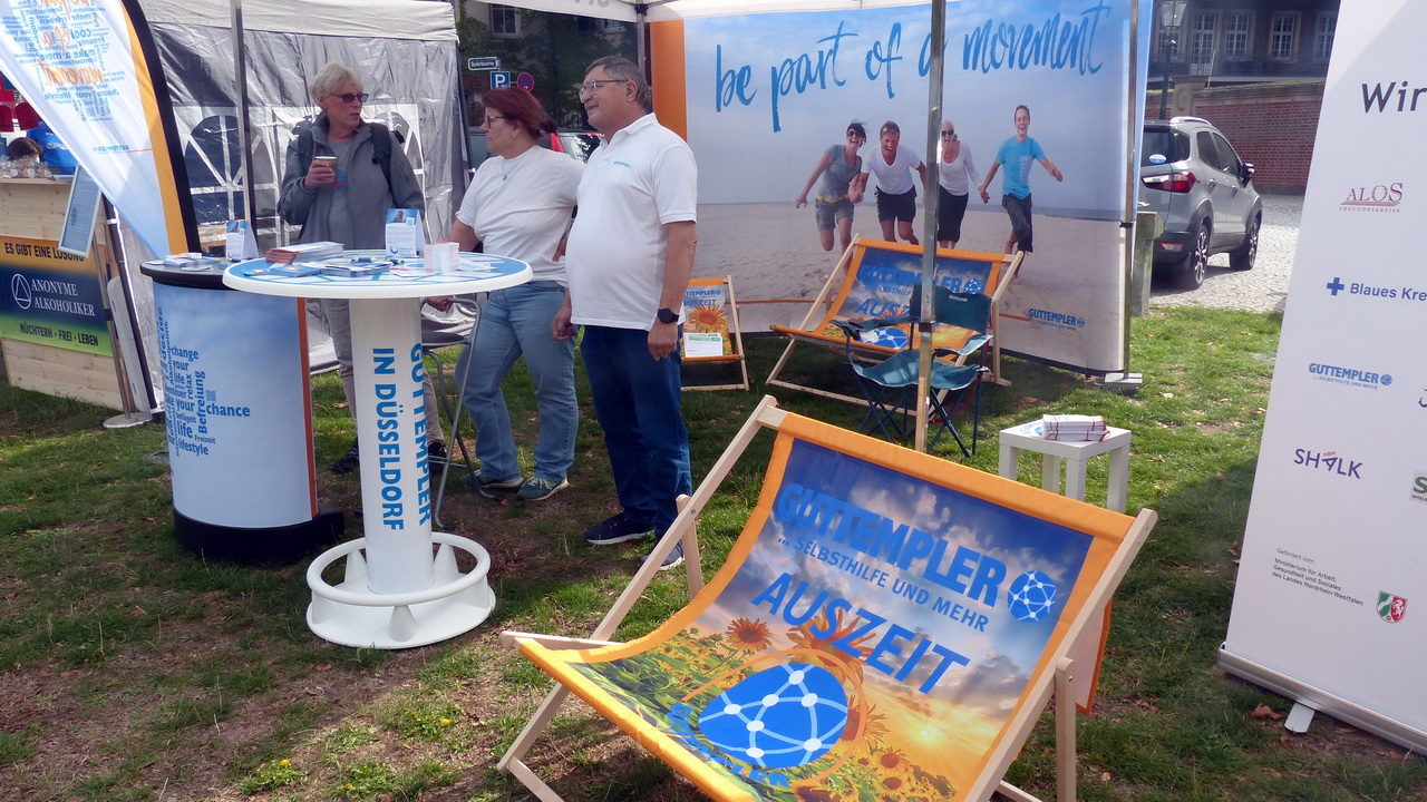Outdoor-Infostand der Guttempler-Selbsthilfegruppe in Düsseldorf: Drei Personen stehen an einem weißen runden Stehtisch, daneben befindet sich ein orangefarbener Liegestuhl mit der Aufschrift 'Guttempler – Selbsthilfe und mehr – Auszeit'. Im Hintergrund sind Werbebanner mit dem Slogan 'be part of a movement' sowie Logos von Kooperationspartnern wie 'Anonyme Alkoholiker' und dem 'Blauen Kreuz' auf einer grasbewachsenen Veranstaltungsfläche sichtbar.