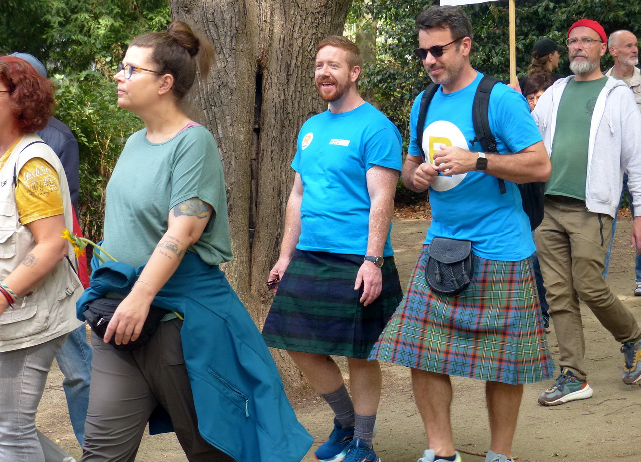 Zwei Männer in blauen T-Shirts und Schottenröcken (Kilts) gehen gemeinsam mit einer Gruppe von Menschen auf einem Weg im Freien. Der Mann rechts trägt eine Sonnenbrille, einen Rucksack und einen bunten Tartan-Kilt, während der Mann links rote Haare und einen dunkleren Kilt trägt. Links im Vordergrund geht eine Frau mit Brille und grünem Shirt.