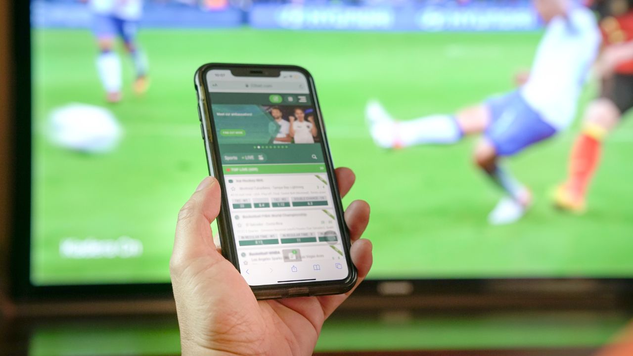 Eine Hand hält ein Smartphone, auf dem eine Sportwetten-Website mit Live-Quoten für Fußballspiele zu sehen ist. Im unscharfen Hintergrund zeigt ein großer Fernseher ein laufendes Fußballspiel auf einem grünen Rasen.