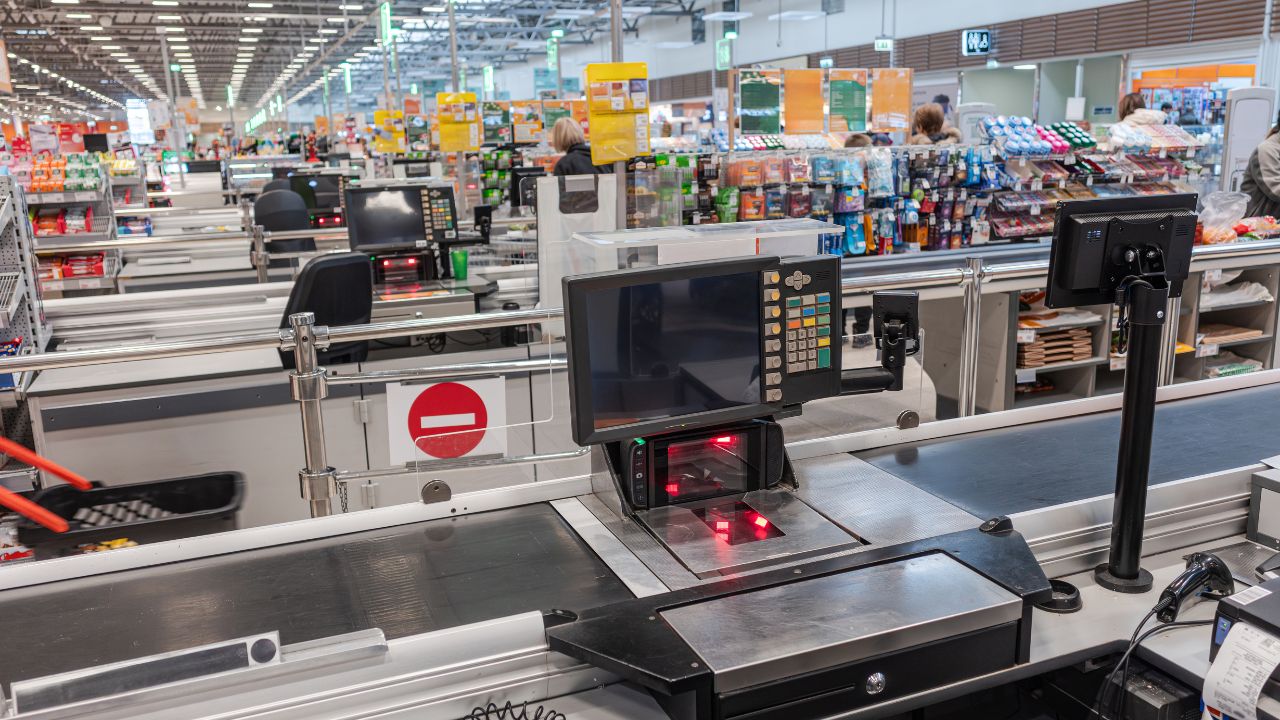Moderne Supermarktkasse mit leerem Förderband, Barcode-Scanner mit rotem Licht und Monitor; im Hintergrund weitere Kassenreihen und Regale eines hellen Lebensmittelgeschäfts.