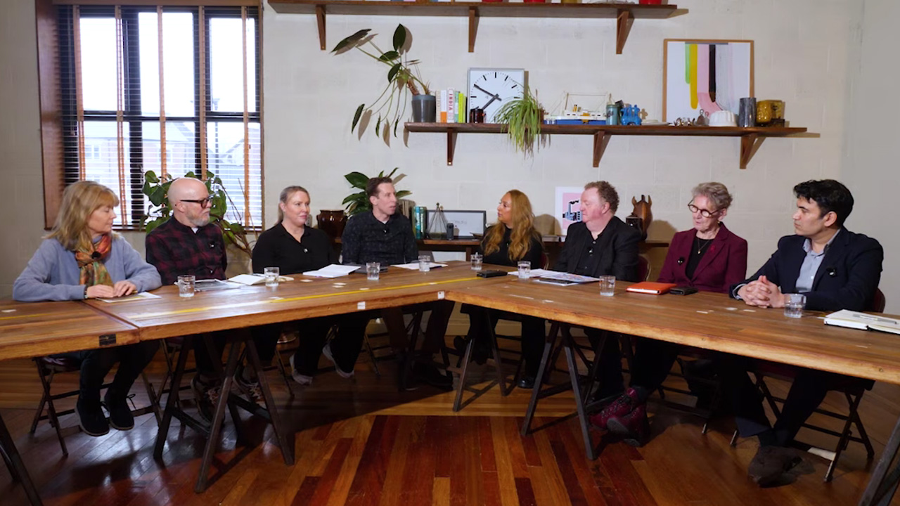 Vier Frauen und vier Männer sitzen an einem U-förmig angeordneten Holztisch in einem kreativen Besprechungsraum und führen eine Diskussionsrunde. Auf dem Tisch stehen Wassergläser und Notizen. Im Hintergrund befinden sich Holzregale mit Pflanzen, Büchern, Kunstwerken und Dekorationsgegenständen sowie ein Fenster mit Jalousien auf der linken Seite. Der Raum hat einen polierten Holzboden und weiße Wände.