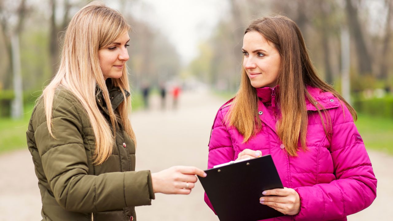 Zwei Frauen in Winterjacken stehen auf einem Parkweg und schauen auf ein Clipboard: Die Blonde in Grün zeigt darauf, die Braunhaarige in Pink hält es fest. Im Hintergrund unscharfe Bäume und Spaziergänger.