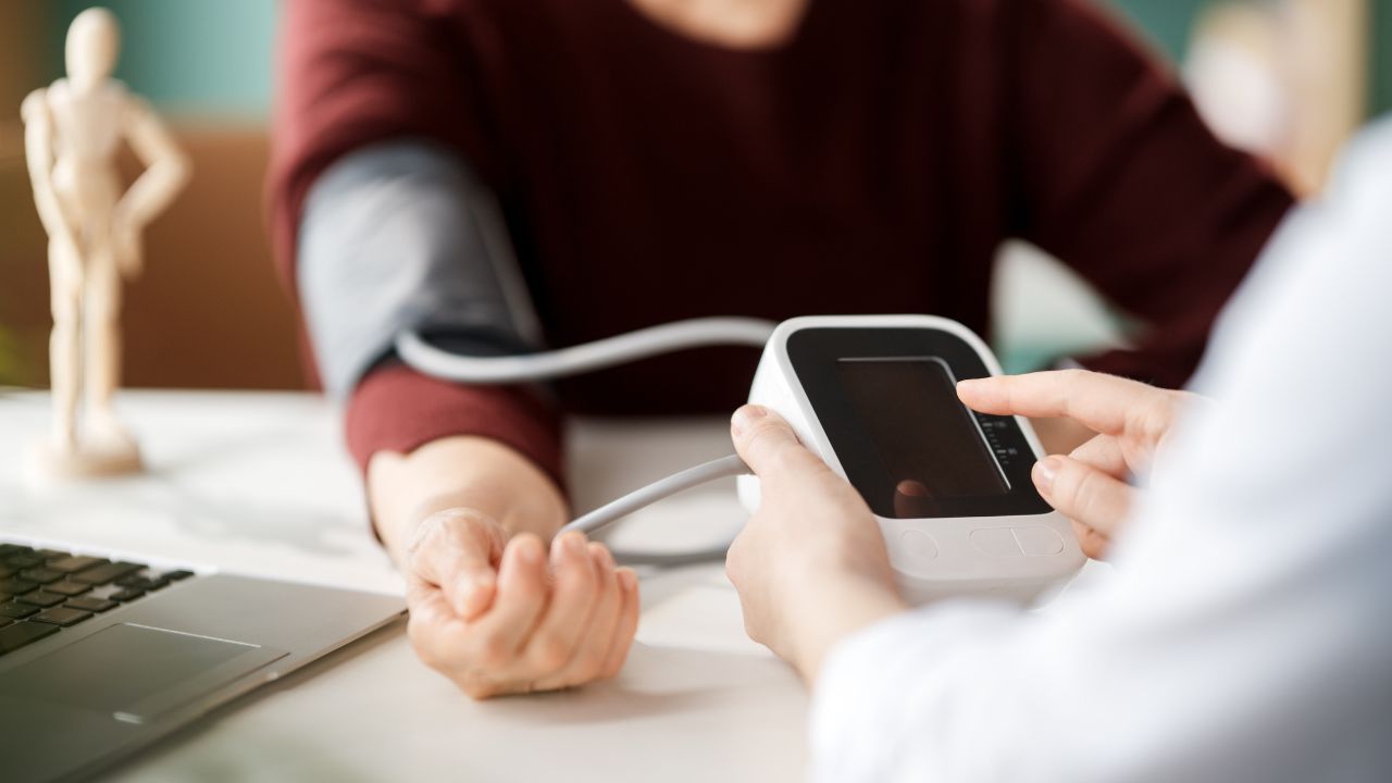 Eine medizinische Fachkraft in weißem Kittel misst mit einem digitalen Blutdruckmessgerät den Blutdruck einer Frau mit rotem Pullover am Tisch. Laptop und Holzfigur im Hintergrund.