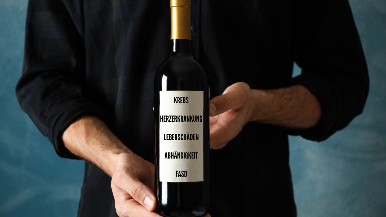 Mann hält eine Weinflasche vor seiner Brust, auf deren Etikett 'Krebs, Herzerkrankung, Leberschäden, Abhängigkeit, FASD' steht.