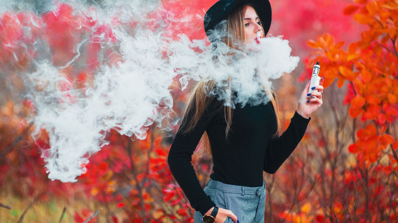 Junges Mädchen mit schwarzem Hut, das im Herbst im Freien ein Vape-Gerät mit einer Wolke aus Dampf verwendet.