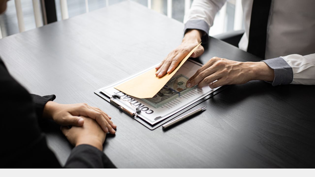 Zu sehen sind die Hände zweier Männer auf einem Schreibtisch. Der eine von ihnen legt Geldscheine zu einem unter einem Umschlag verborgenen Vertrag.