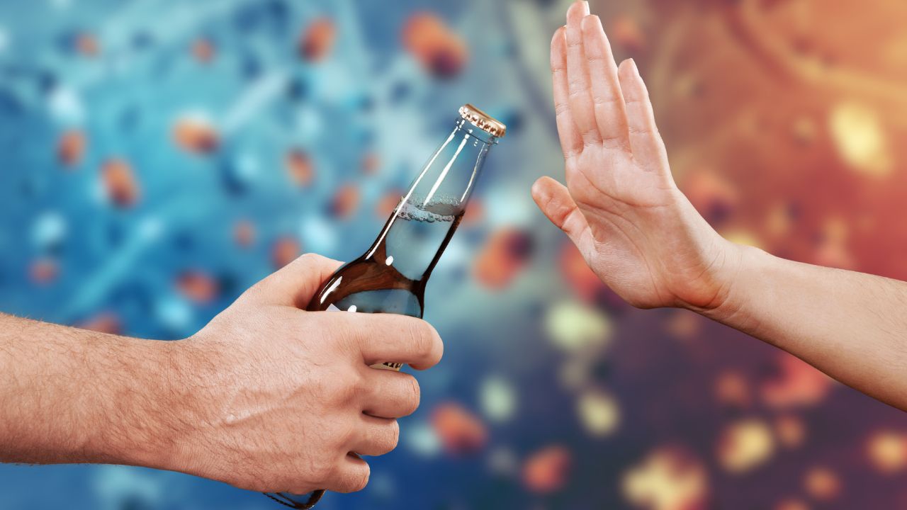 Eine Hand reicht eine Bierflasche an, während eine andere Hand mit abwehrender Geste signalisiert, das Getränk abzulehnen. Im Hintergrund ist eine unscharfe, farbige Darstellung von Krebszellen zu sehen.