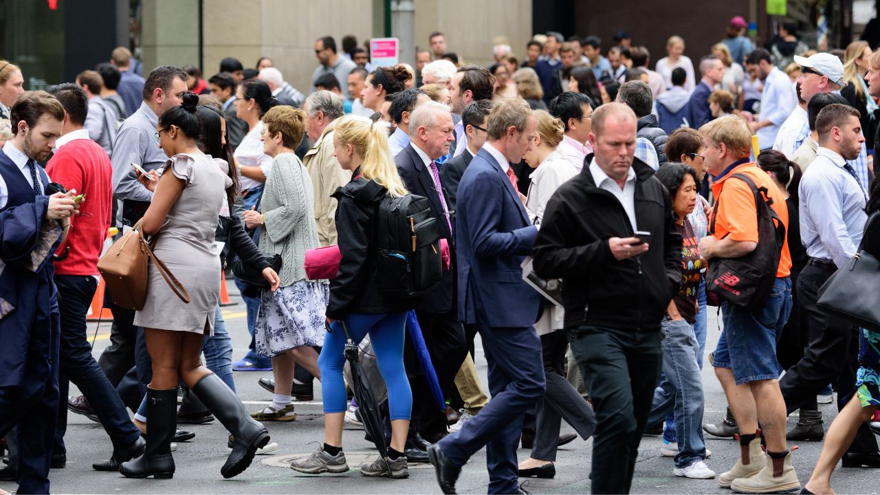 Eine dichte Gruppe von Fußgängern und Pendlern unterschiedlichen Alters auf einem belebten Stadtweg, von denen viele auf ihre Smartphones blicken.