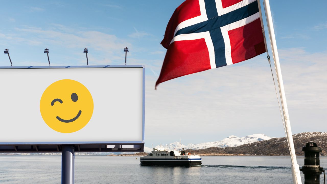 Leere Werbetafel mit einem zwinkernden Smiley vor norwegischer Fjordlandschaft mit norwegischer Flagge im Vordergrund.