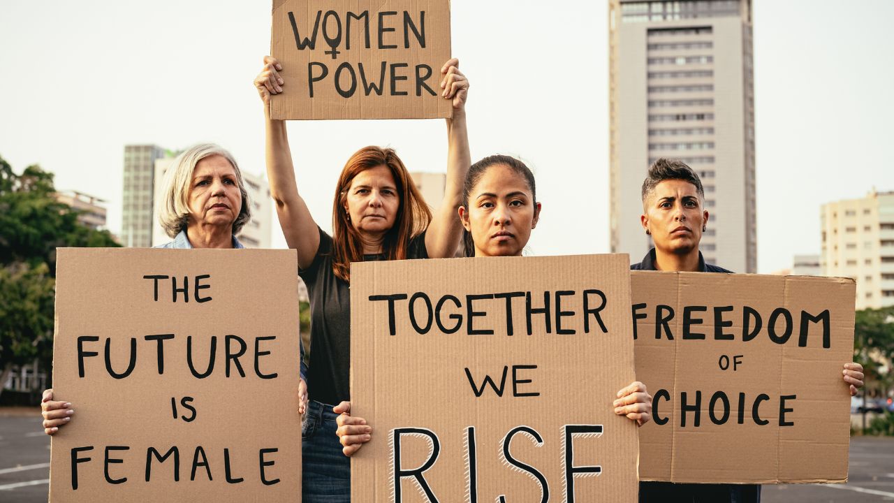 Vier Personen unterschiedlichen Alters und verschiedener Herkunft stehen bei einer Protestaktion im Freien nebeneinander und halten handgeschriebene Pappschilder hoch. Die Schilder tragen feministische Botschaften: 'THE FUTURE IS FEMALE', 'TOGETHER WE RISE', 'WOMEN POWER' (mit Venus-Symbol) und 'FREEDOM OF CHOICE'. Die Demonstrierenden blicken ernst und entschlossen in die Kamera. Im Hintergrund sind unscharf Hochhäuser und Stadtbäume zu sehen.