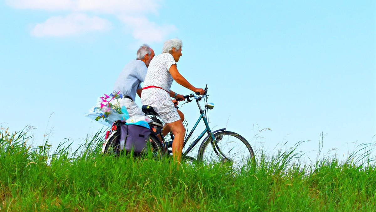Zwei radfahrende Senioren hinter grünem Gras und vor blauem Himmel