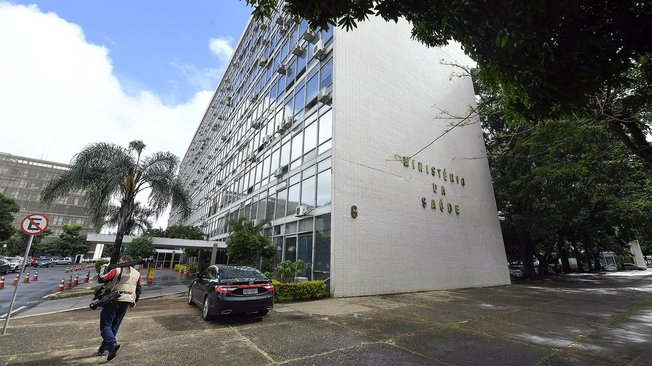 Das moderne Hauptgebäude des brasilianischen Gesundheitsministeriums (Ministério da Saúde) an einem sonnigen Tag, mit geparktem schwarzem Auto und Passant im Vordergrund.