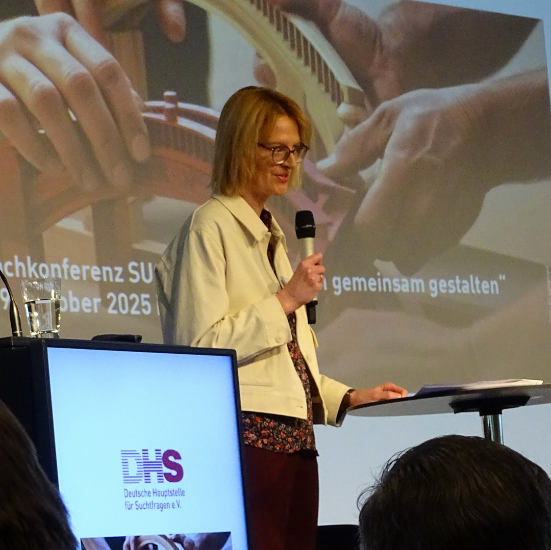 Christina Rummel steht an einem Rednerpult mit DHS-Logo, im Hintergrund ist eine Leinwand mit dem Titel der Veranstaltung zu sehen.